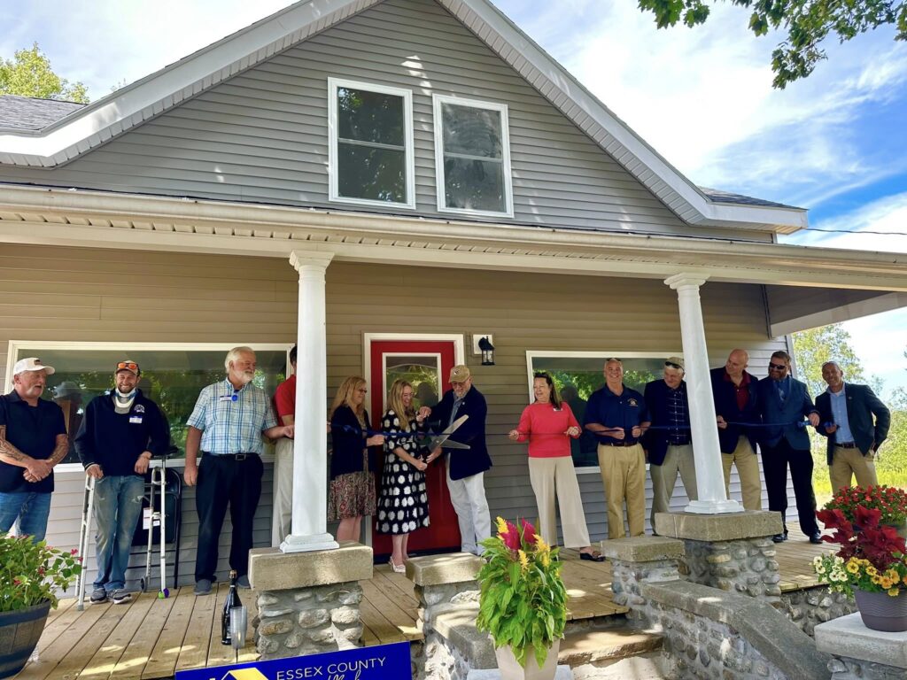 men and women stand on the porch of newly renovated home with red door and cut blue ribbon with large scissors