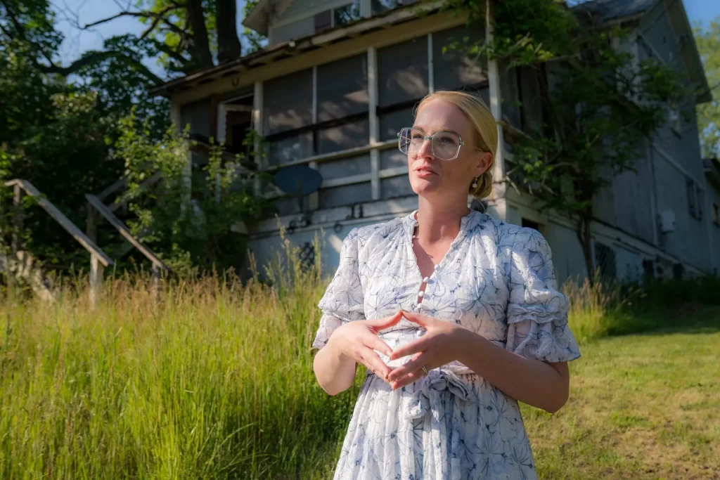 Land Bank CEO, Nicole Justice Green, blonde woman with glasses stands in front of a dilapidated home to be rehabilitated